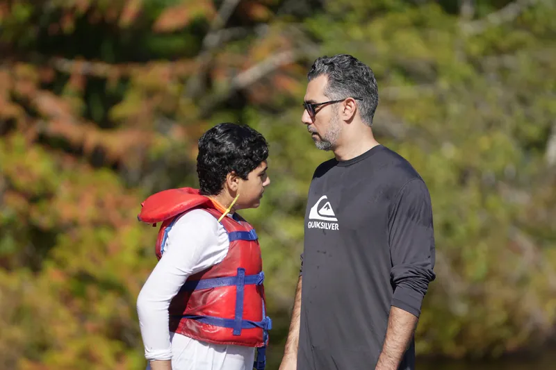 A man and a boy in a red life jacket stand face to face talking at the edge of a lake, with autumn trees behind them.