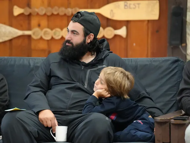 A young boy rests his chin on his hand as he leans against a bearded man on a sofa during a session, with wooden canoe paddles on the wall behind.