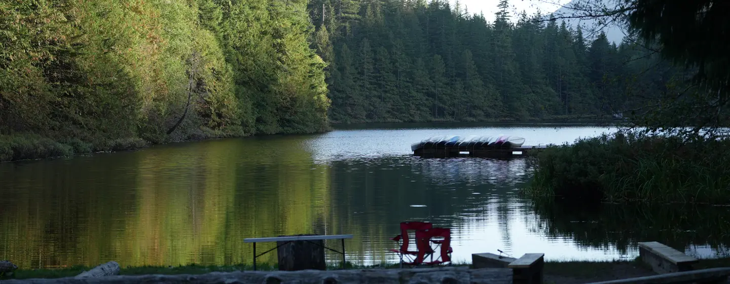 Evening light on a calm forest lake with a dock of canoes, a picnic table and red camp chairs on the shore.